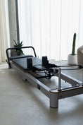 Load image into Gallery viewer, Pilates reformer machine in a room with large windows and plants.
